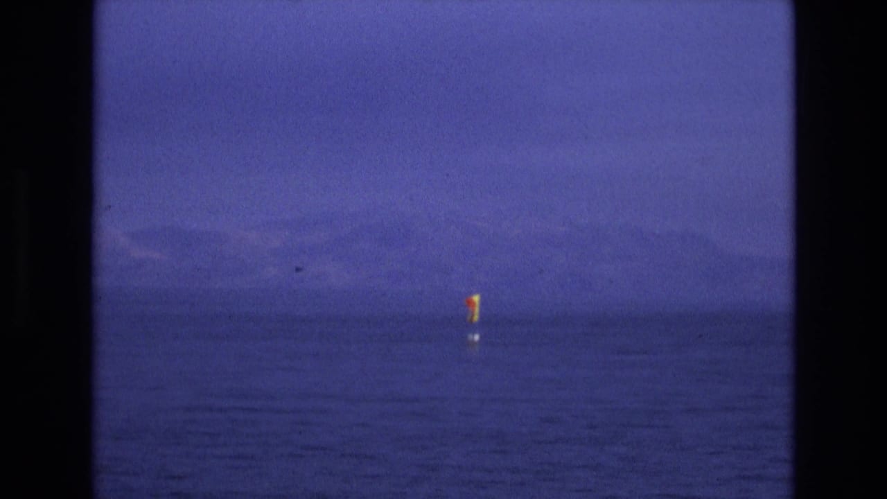 Distant red and yellow flag on a buoy in calm blue water with hazy mountains behind it.