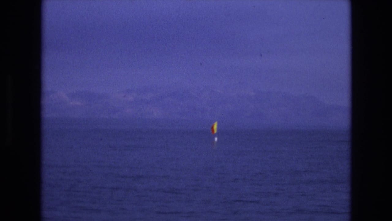 Distant buoy-mounted flag on calm water in the supplied 1973 Salt Lake City, Utah home-movie footage, framed by dark film edges and a hazy mountain horizon.