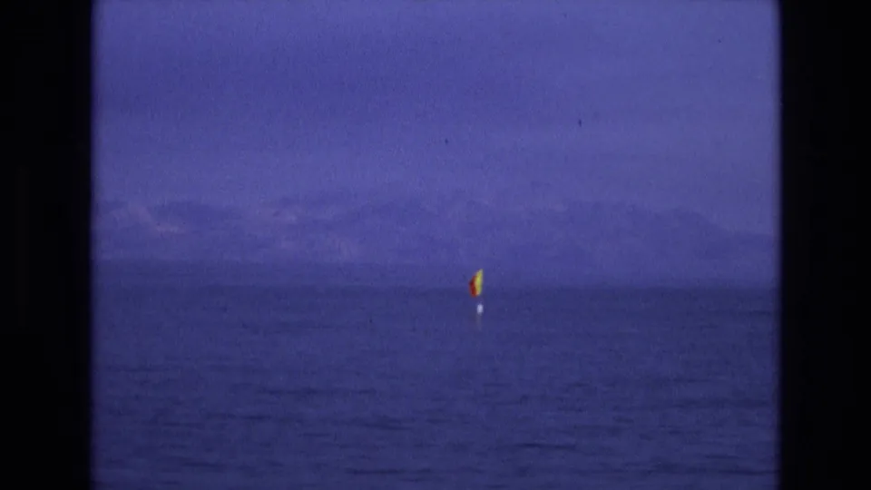 Distant buoy-mounted flag on calm water in the supplied 1973 Salt Lake City, Utah home-movie footage, framed by dark film edges and a hazy mountain horizon.