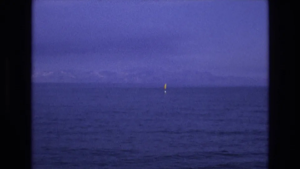 1973 8mm home-movie frame from the supplied Salt Lake City, Utah metadata showing calm blue water, a low distant horizon, and a small yellow buoy-like marker seen through dark vertical framing.