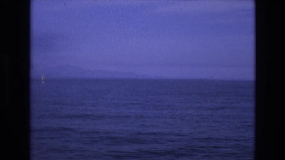 1973 8mm home-movie frame from the supplied Salt Lake City, Utah metadata showing calm blue water, a faint horizon, and a small yellow marker framed by dark vertical edges.