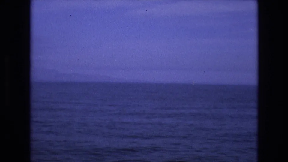 1970s 8mm home-movie frame showing a dark blue ocean horizon with gentle waves under a low, dusky sky, viewed through heavy black vertical framing.