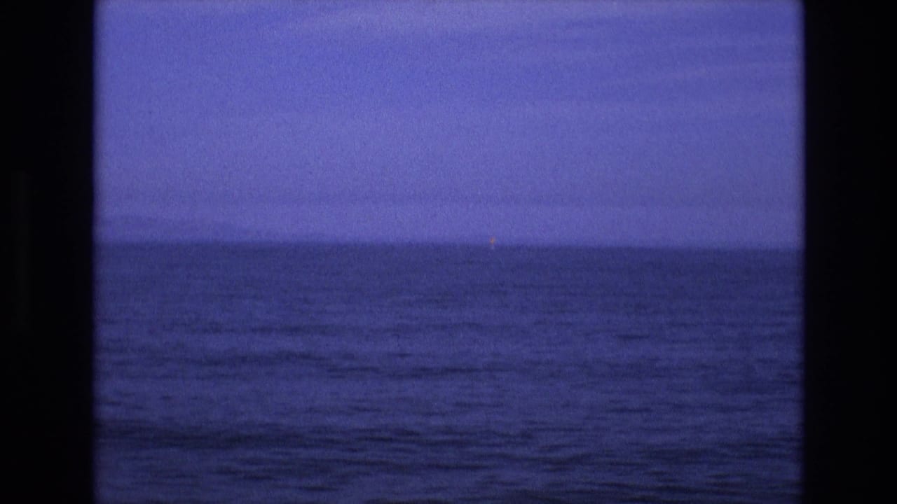1970s 8mm home-movie frame of rolling blue water and a dim horizon, with a small distant light visible near the right side of the seascape.