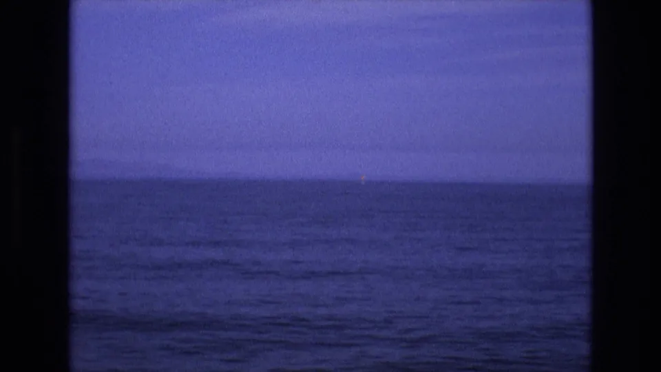 1970s 8mm home-movie frame of rolling blue water and a dim horizon, with a small distant light visible near the right side of the seascape.