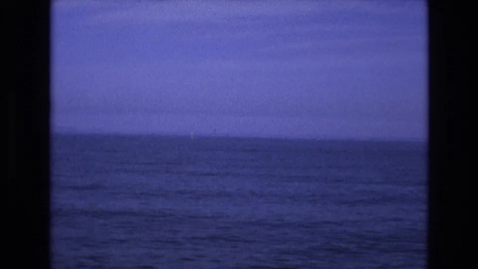 1970s 8mm home-movie frame showing a softly focused ocean surface, a low horizon, and a pale blue-purple sky band within dark vertical framing.
