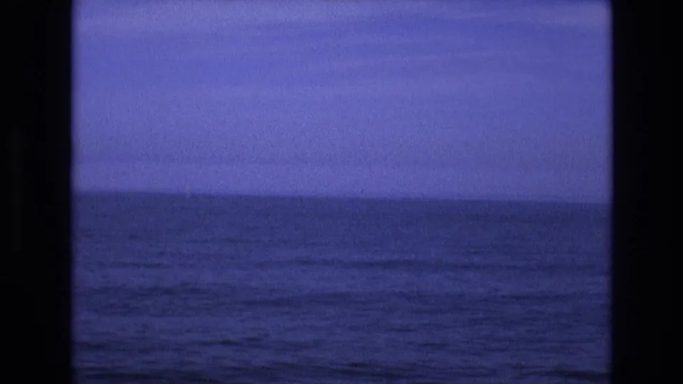 1970s 8mm home-movie frame with dark rolling ocean water, a muted horizon, and black vertical framing that suggests filming through a window or structural opening.