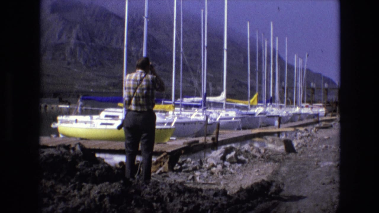 Man standing on a rocky shore with binoculars near sailboats and tall masts.