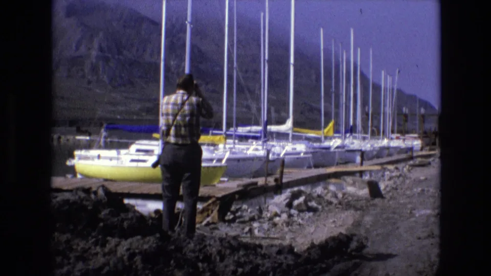 1973 8mm Salt Lake City home-movie frame of a man in a plaid shirt looking through binoculars beside a marina with sailboats and mountains behind it.