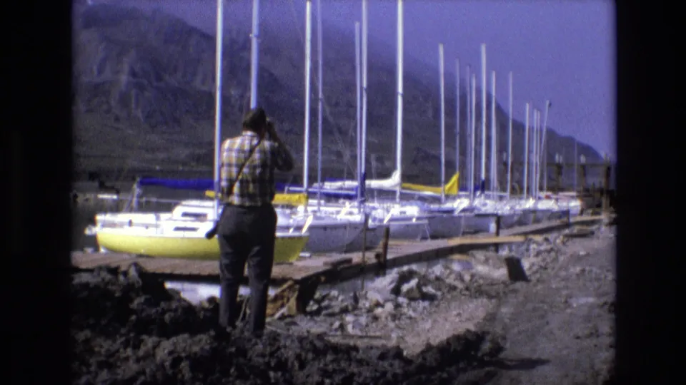 1973 8mm Salt Lake City home-movie frame showing a man from behind on rocky ground near a yellow sailboat, white masts, a dock, and mountain scenery.