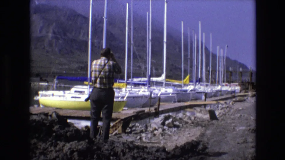 1973 8mm Salt Lake City home-movie frame of a man observing a marina through binoculars, with a yellow boat, clustered masts, shoreline rocks, and mountains visible.