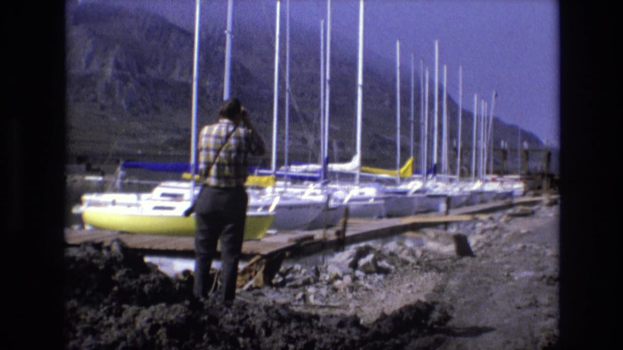 In 1973 Salt Lake City area home-movie footage, a man in a plaid shirt stands on rocky ground looking through binoculars toward docked sailboats below rugged mountains.