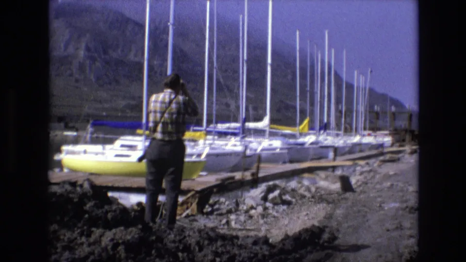 In 1973 Salt Lake City area home-movie footage, a man in a plaid shirt stands on rocky ground looking through binoculars toward docked sailboats below rugged mountains.