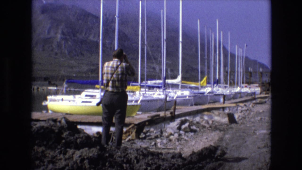 Man stands facing a marina filled with sailboats and tall masts near mountains.