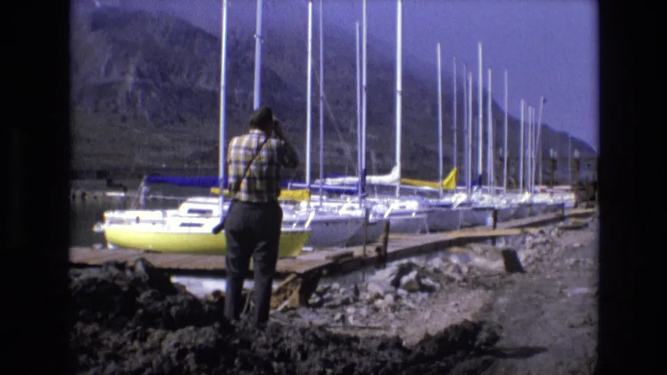 A lone adult man faces away from the camera toward a marina of sailboats and masts, with rocky terrain and mountain slopes visible in the Salt Lake City area footage.