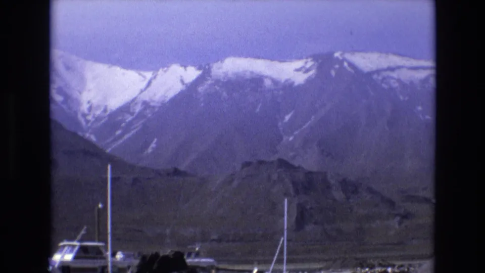 Snow-streaked mountains dominate the frame above a marina edge with a few boat masts and a rocky shoreline, filmed in soft handheld 8mm style.