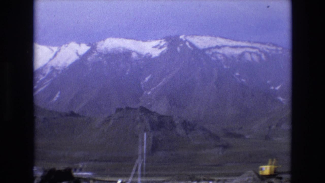 1973 Salt Lake City 8mm frame showing snow-streaked mountains under a hazy sky, rocky foreground terrain, white poles, and a small yellow excavator near the lower edge.