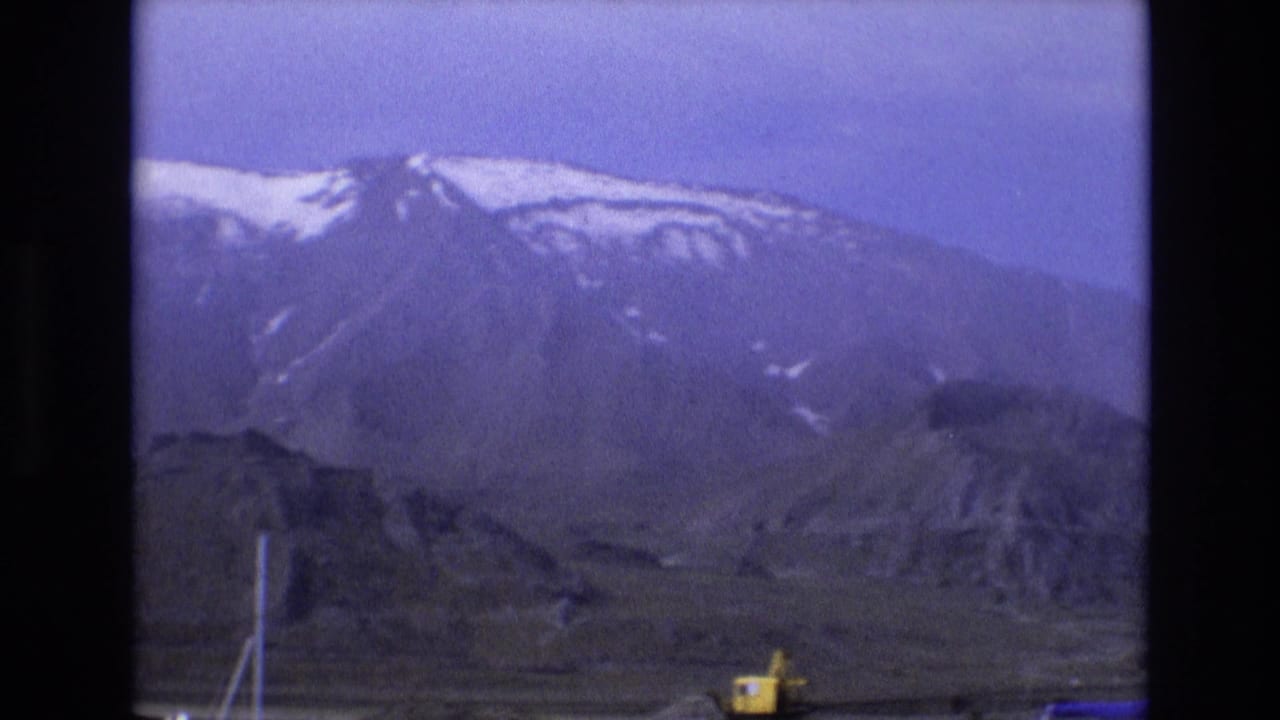 Distant snow-capped mountains with rocky terrain and a yellow excavator near the bottom edge.