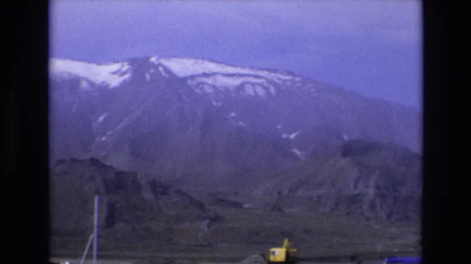 1973 Salt Lake City frame with a broad mountain ridge, patches of snow, barren lower terrain, and a small yellow excavator visible near the bottom of the shot.