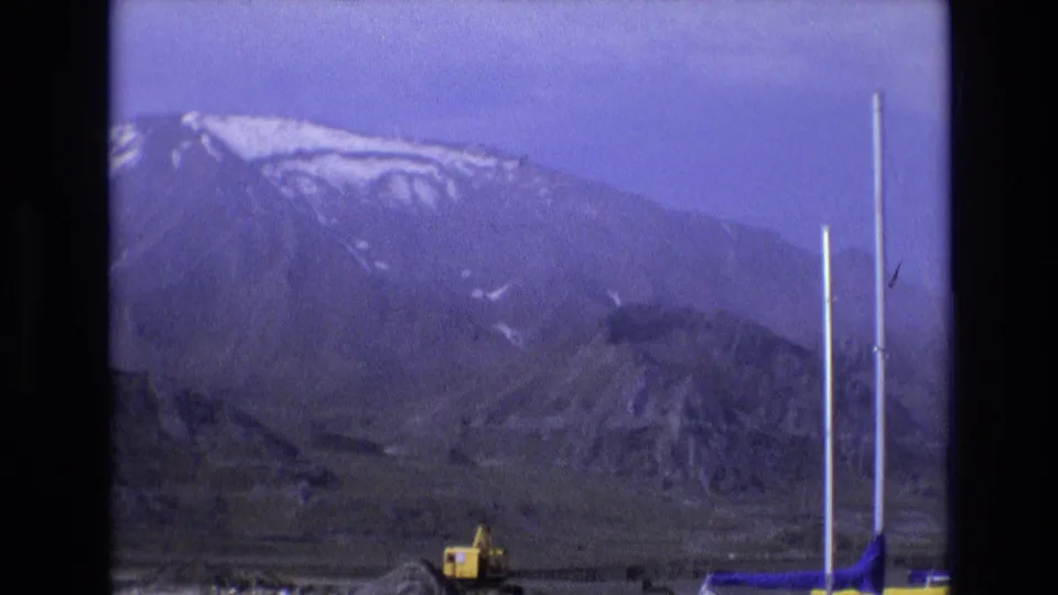 1973 Salt Lake City 8mm frame showing a hazy mountain slope, rocky lower ground, several white poles or masts, and a yellow excavator below the ridge.