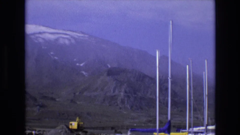 1973 Salt Lake City frame with multiple white vertical poles or masts in the foreground, a yellow excavator near the bottom, and snow-marked mountains in the distance.