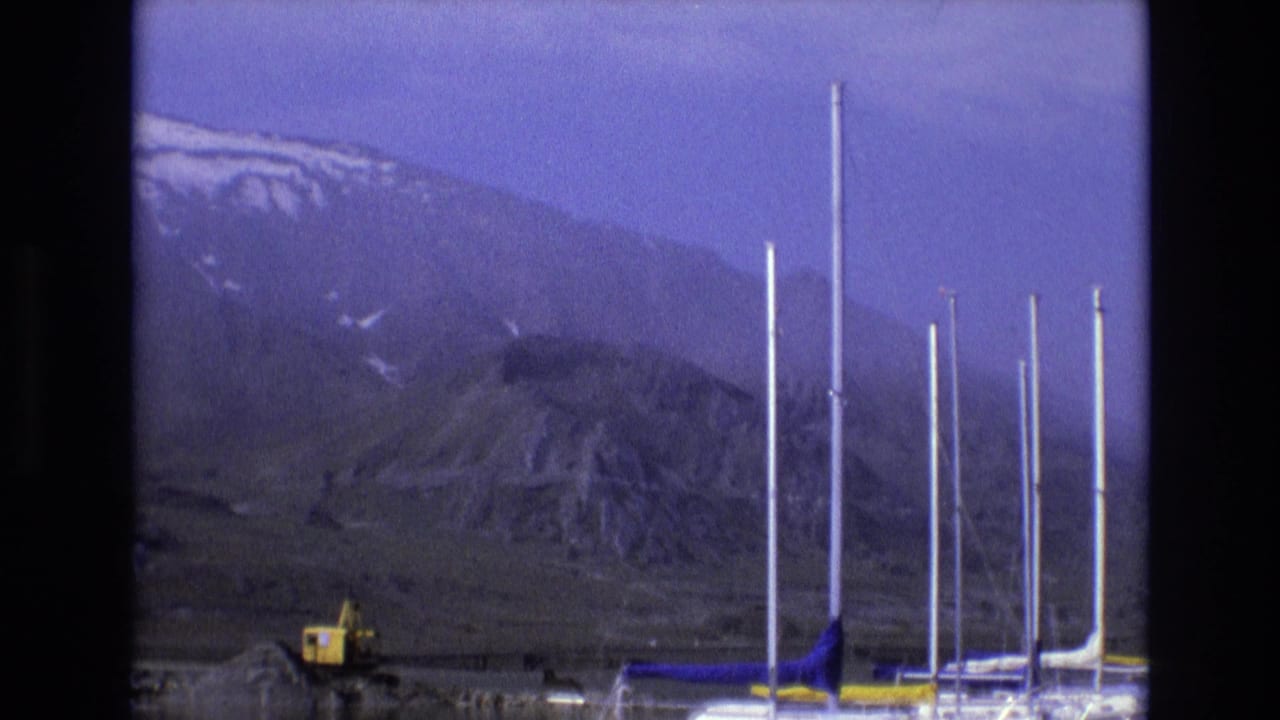 1973 Salt Lake City area 8mm frame showing rugged snow-streaked mountains, a yellow excavator, and several tall masts or poles near the lower edge of the scene.