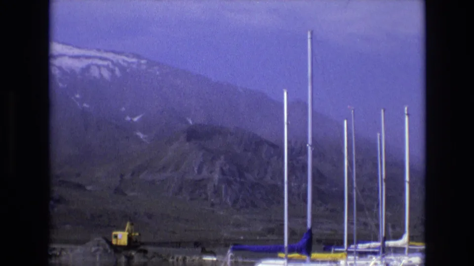 1973 Salt Lake City area 8mm frame showing rugged snow-streaked mountains, a yellow excavator, and several tall masts or poles near the lower edge of the scene.