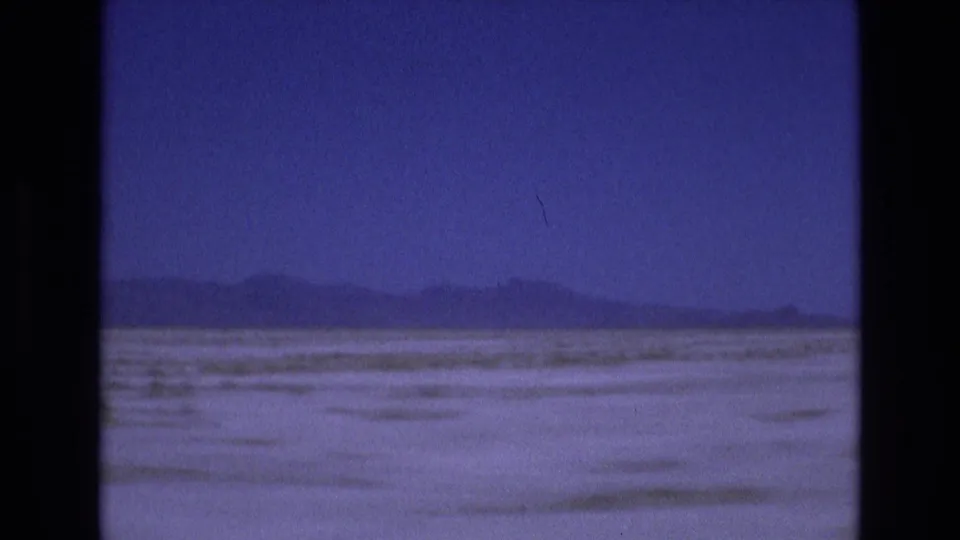 1973 Utah 8mm frame showing a broad pale salt flat or dry lakebed with low mountains on the horizon beneath a blue-purple sky.