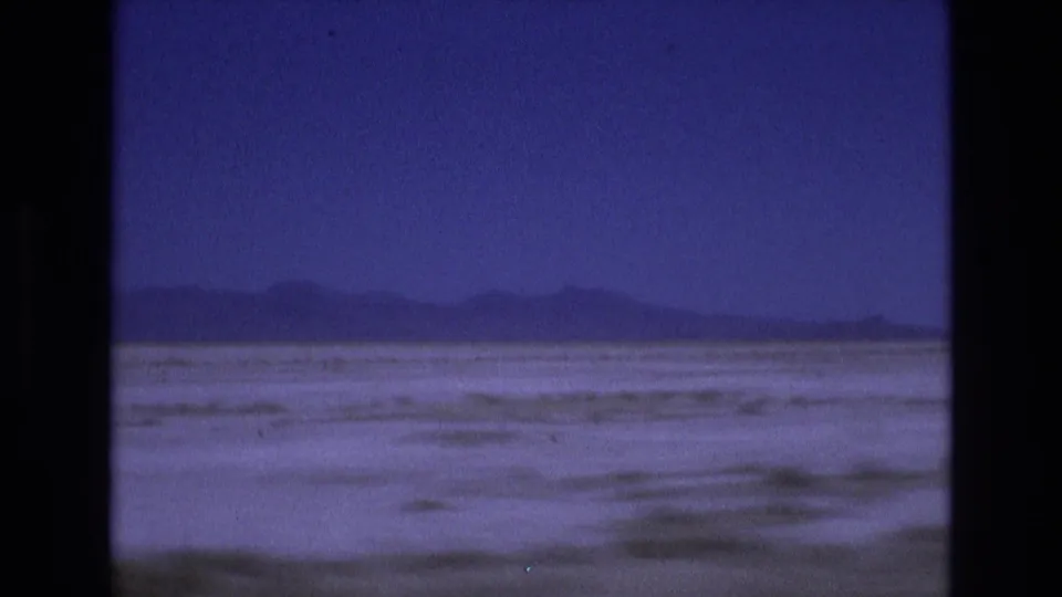 1973 Utah 8mm frame showing textured salt-flat ground stretching toward a low mountain horizon under a deep blue-purple sky.