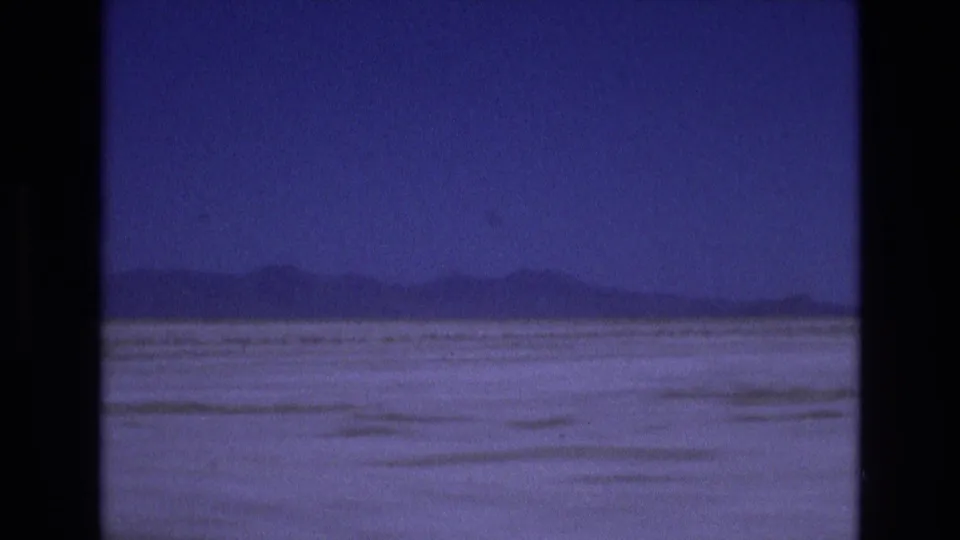 1973 Salt Lake City area home-movie frame showing a pale flat basin or salt-flat landscape with low mountain silhouettes beneath a deep blue sky.