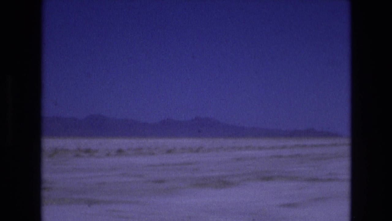 1973 Utah home-movie frame with distant mountain forms stretching across a flat pale foreground, seen through soft blue-purple 8mm color.