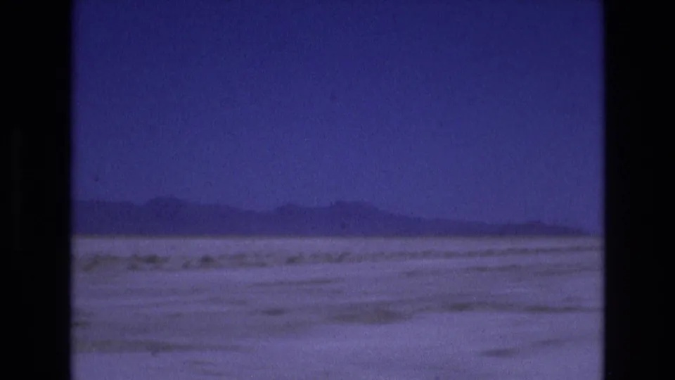 1973 Utah home-movie frame with distant mountain forms stretching across a flat pale foreground, seen through soft blue-purple 8mm color.