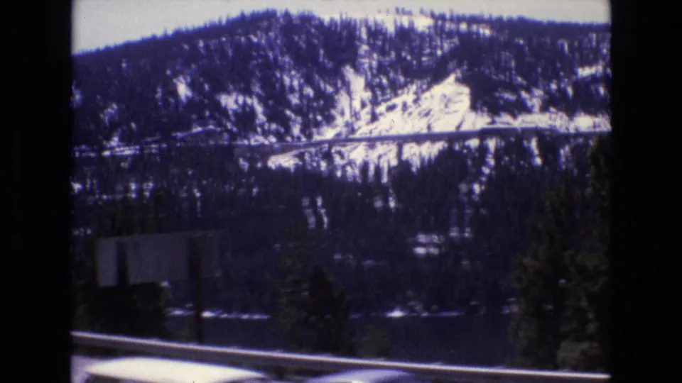 1973 8mm travel footage appears to show a snowy forested mountain slope, dark lake water, roadside guardrail, and trees filmed from a moving vehicle in California.
