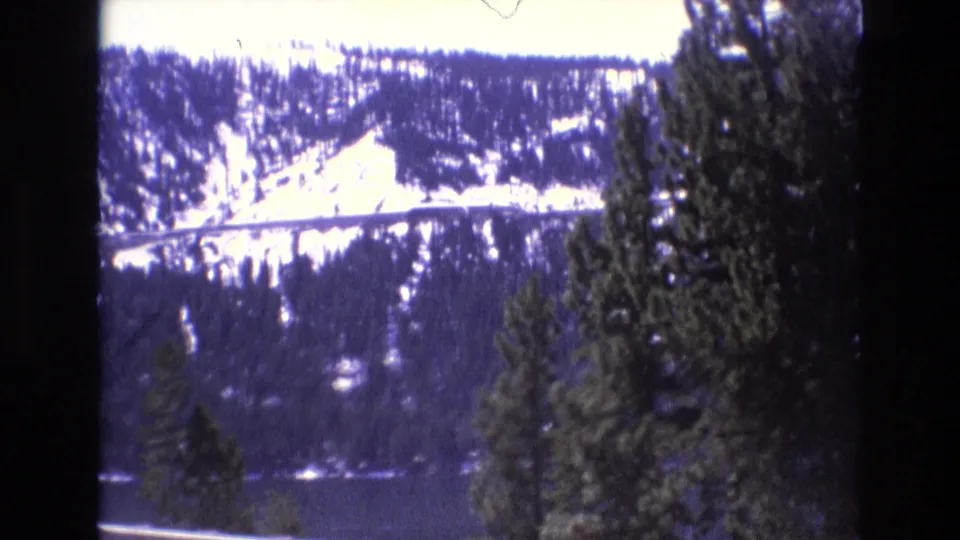 A later 1973 8mm frame shows a snow-covered mountain hillside, dark lake, evergreen trees, and a roadside vantage point with strong contrast and film softness.