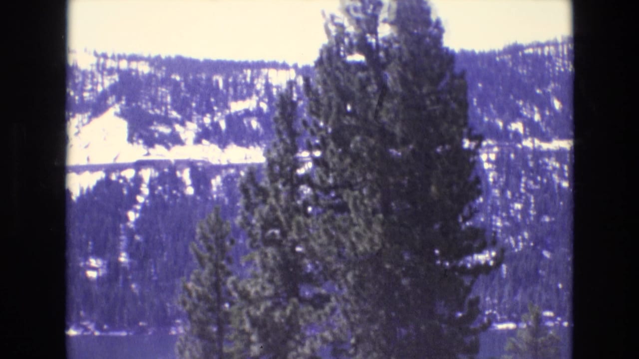 1970s 8mm frame cataloged to California showing evergreen trees, a dark lake, and snow-patched mountain slopes under a bright sky.