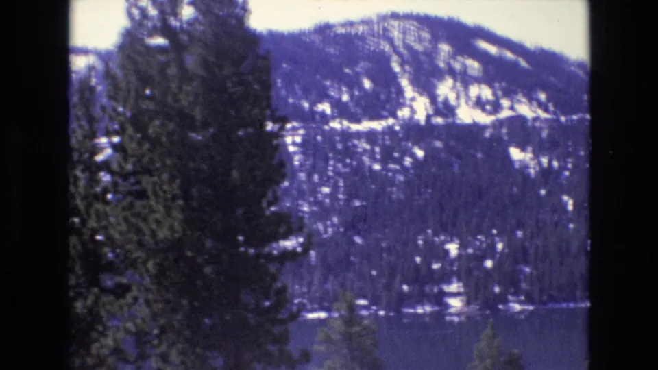 1970s 8mm frame cataloged to California with tall conifers in the foreground, a calm lake, and snow-streaked mountain terrain beyond.