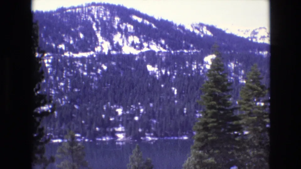 1970s 8mm frame cataloged to California showing a broad lake, evergreen foreground trees, and snow-patched mountains in soft home-movie color.