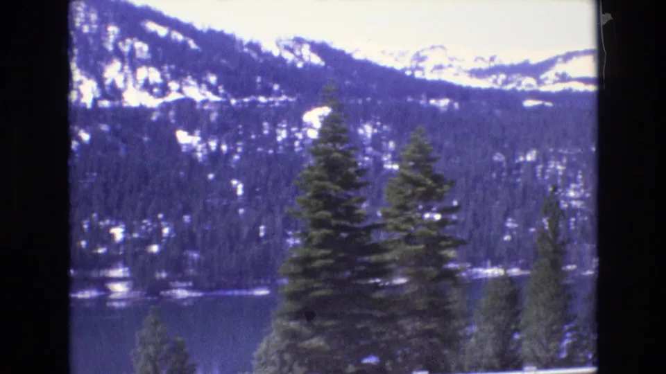 1970s 8mm frame cataloged to California with evergreen trees overlooking a dark lake and snow-streaked mountains in the distance.