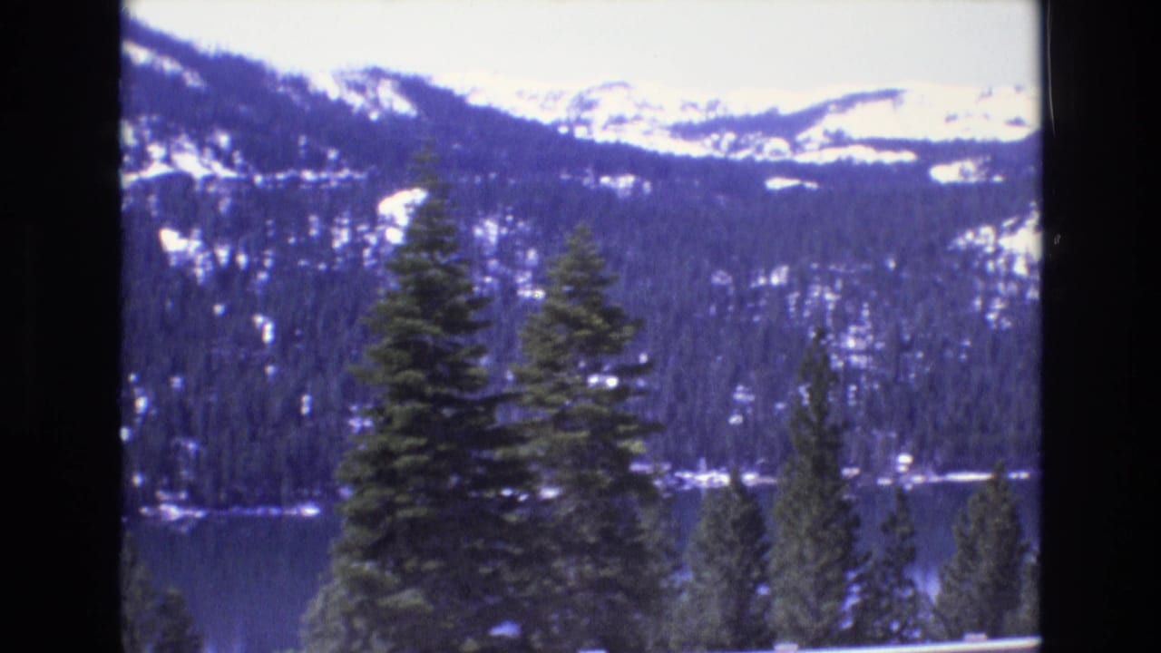 1973 8mm home-movie frame showing snow-capped ridges, dense evergreens, and a lake from an elevated scenic overlook.