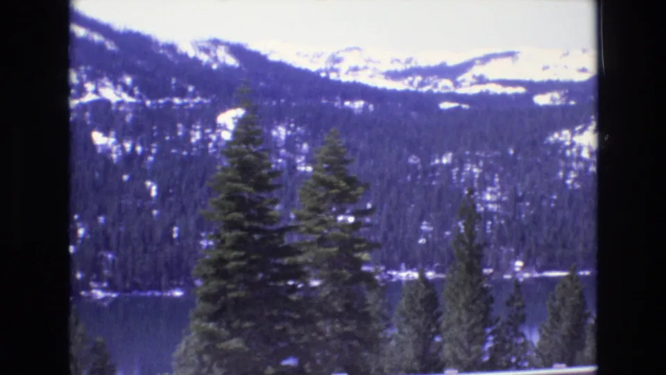 1973 8mm home-movie frame showing snow-capped ridges, dense evergreens, and a lake from an elevated scenic overlook.