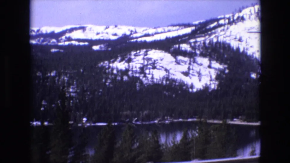 1973 8mm home-movie frame showing a snow-dusted mountain slope, forested lakeshore, and dark reflective water.