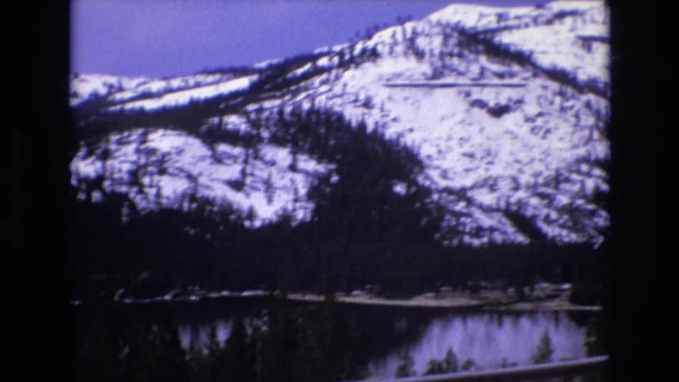 1973 8mm scenic frame of a broad snow-streaked mountainside, evergreen forest, and lake viewed from an overlook.