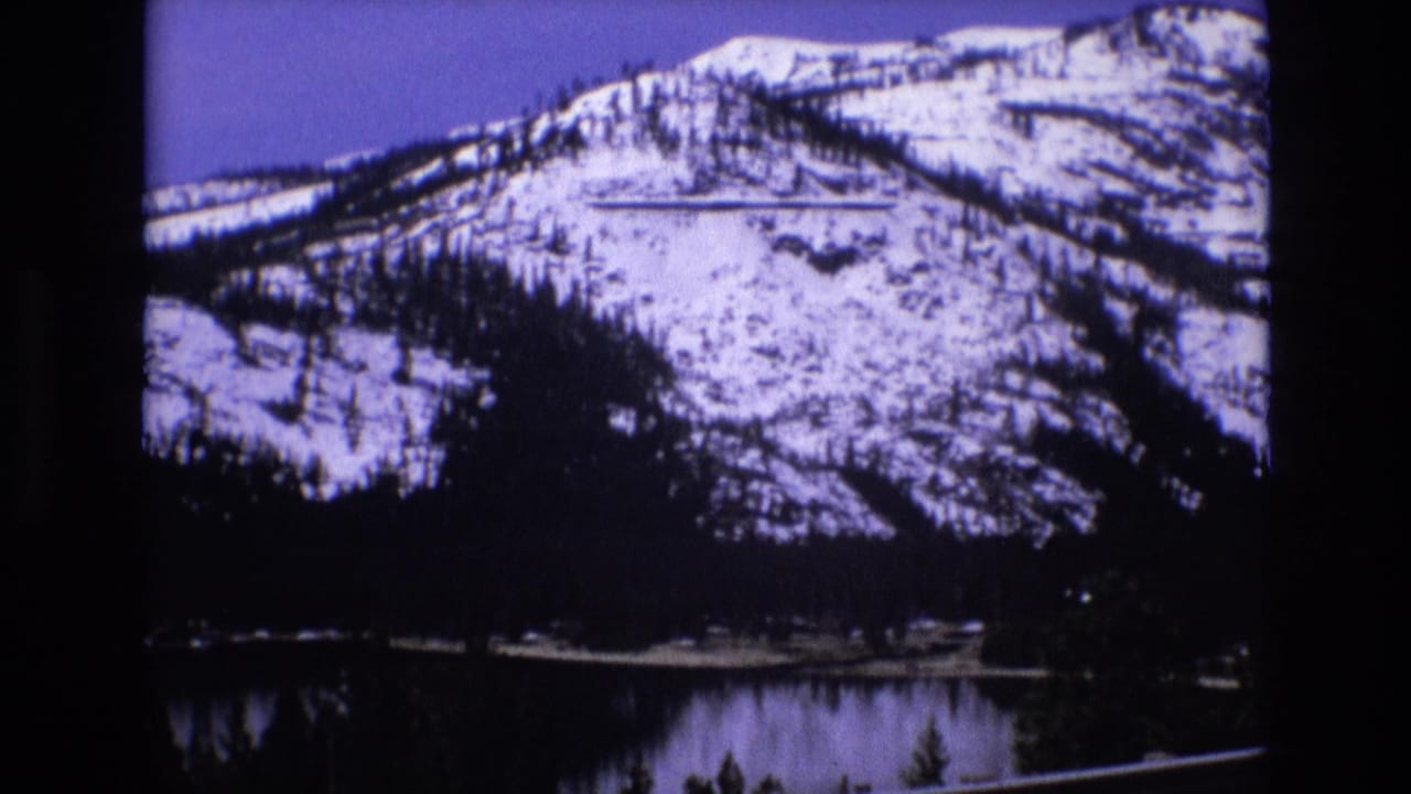 Snow-covered mountain above a dark lake with evergreen trees in the foreground.