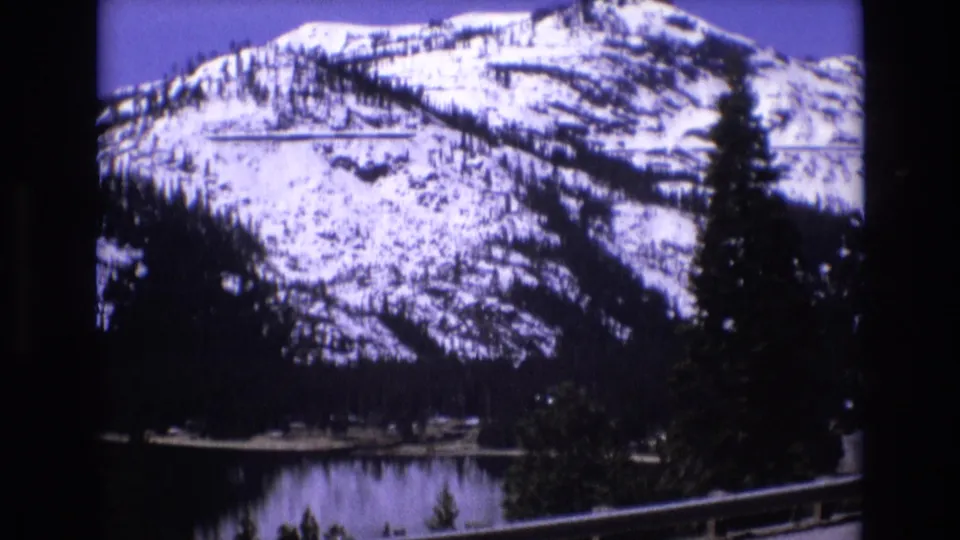 1973 8mm travel frame with a snow-covered mountain, lake, tall evergreen, and dark foreground trees as the camera moves along a road.
