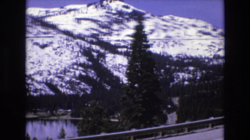 1973 8mm home-movie frame showing a roadside guardrail, lake, evergreen tree, and snow-covered mountain slope in a winter travel scene.