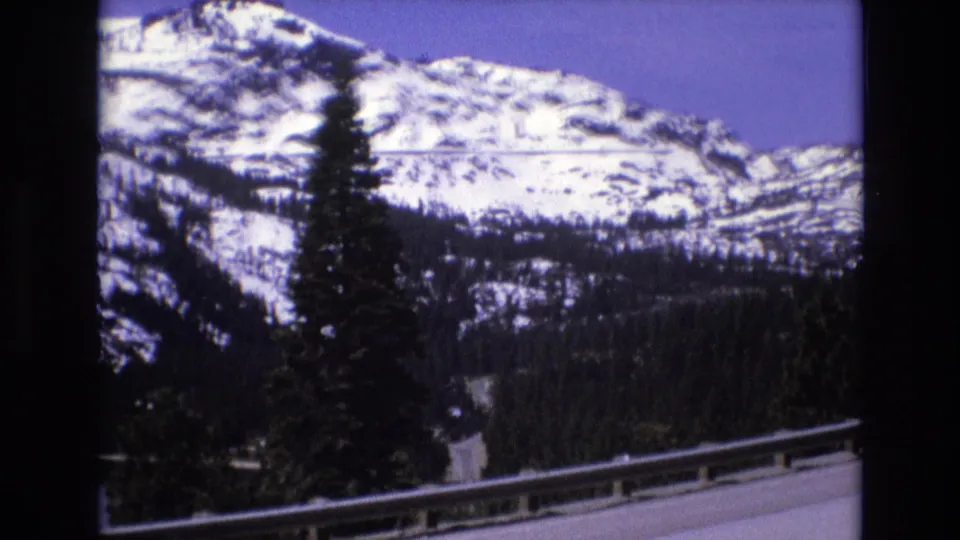 1973 8mm travel frame with a guardrail crossing the foreground, evergreens and lake below, and a snowy mountain ridge beyond.