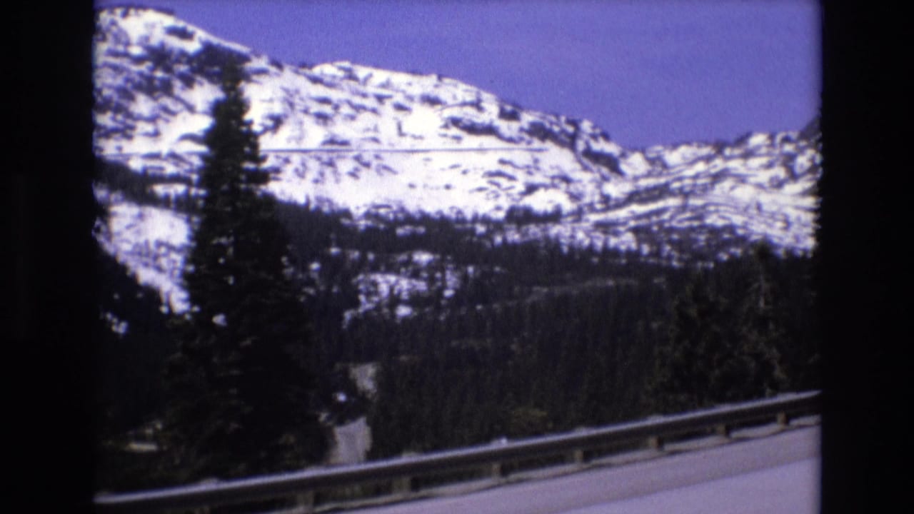1973 8mm frame of a California mountain highway scene, with snow-capped peaks, evergreen trees, and a roadside guardrail seen from a moving vehicle.