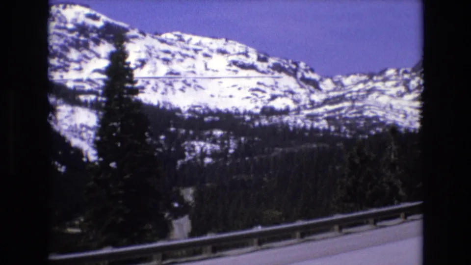 1973 8mm frame of a California mountain highway scene, with snow-capped peaks, evergreen trees, and a roadside guardrail seen from a moving vehicle.