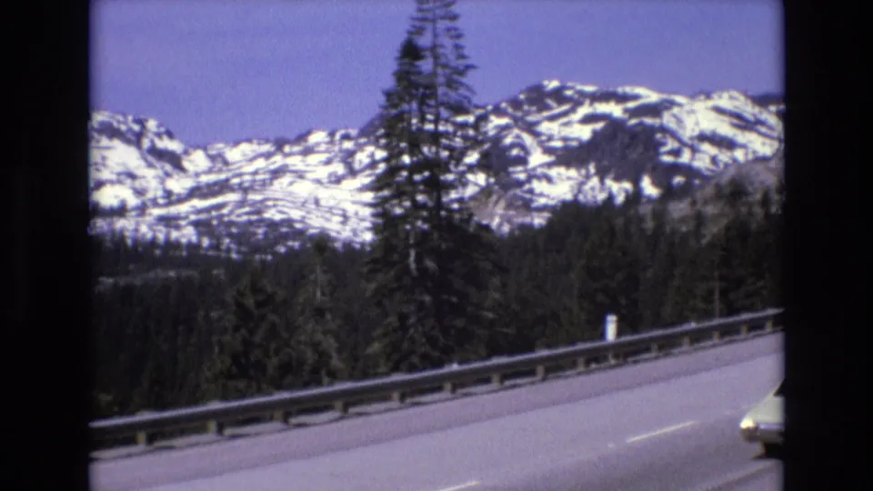 1970s home-movie frame from a mountain highway, showing the road edge, guardrail, conifer trees, and a snow-streaked ridge in the distance.
