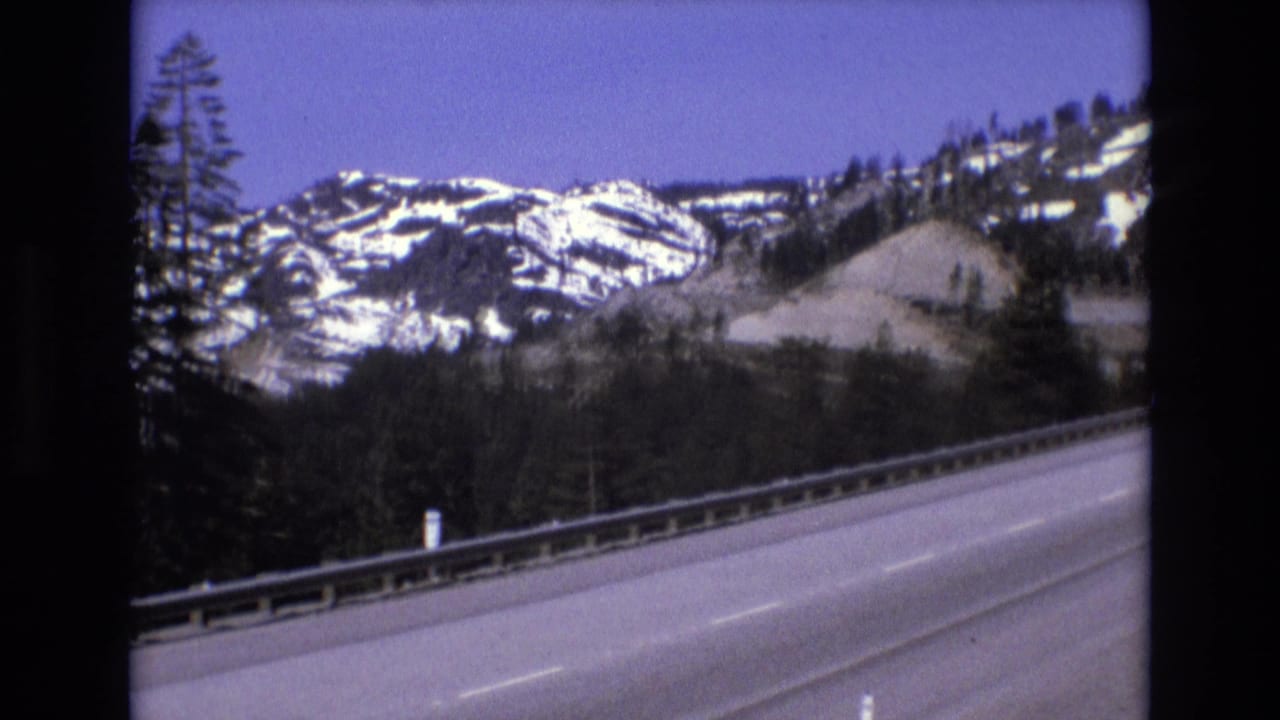 1973 8mm road-travel frame showing a paved highway edge with a metal guardrail, evergreen forest, and snow-dusted mountain slopes in the supplied California context.