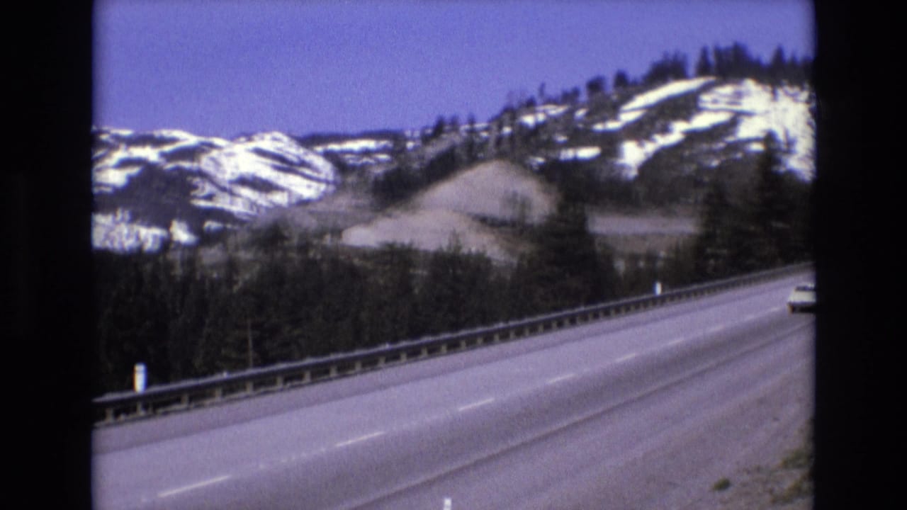 Curving highway with snowy wooded hills and a partial vehicle at the road edge.